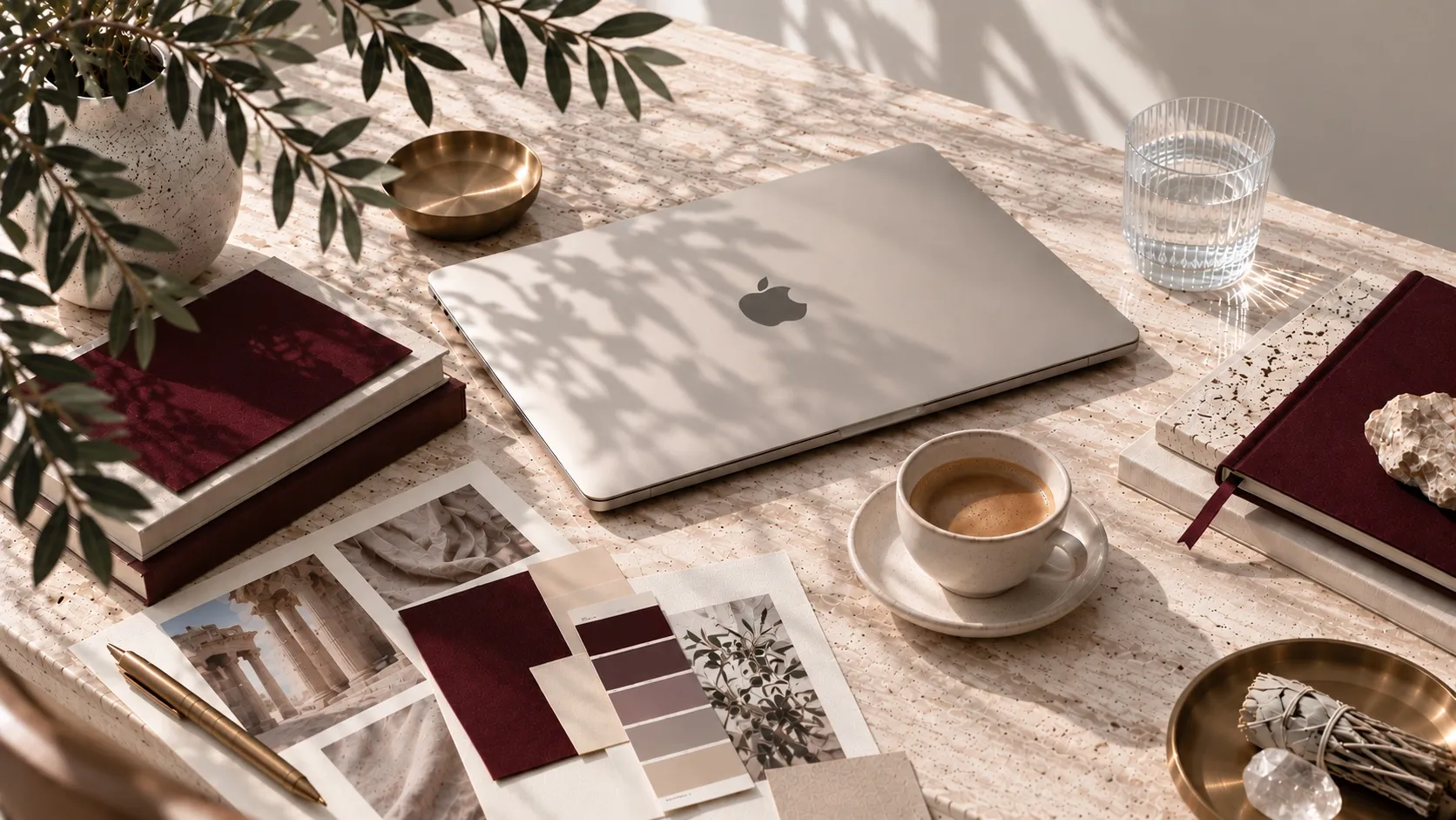 Escritorio con portátil, café, muestras de color y libretas borgoña bajo luz natural